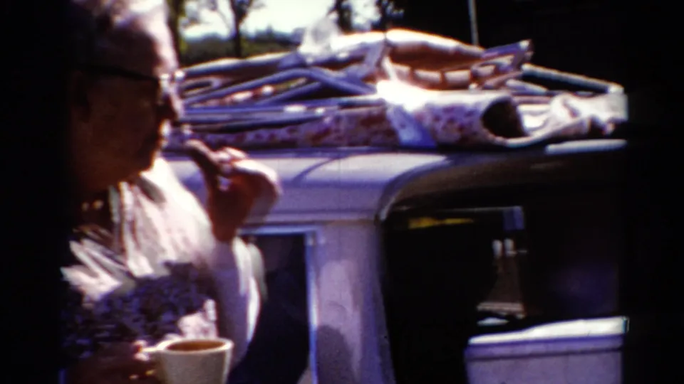 A closer handheld frame shows the woman beside the car, cup in hand, with roof luggage and dark car interior visible.