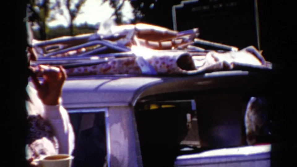 The frame catches the woman partly at the edge as the camera favors the packed car roof and roadside setting.