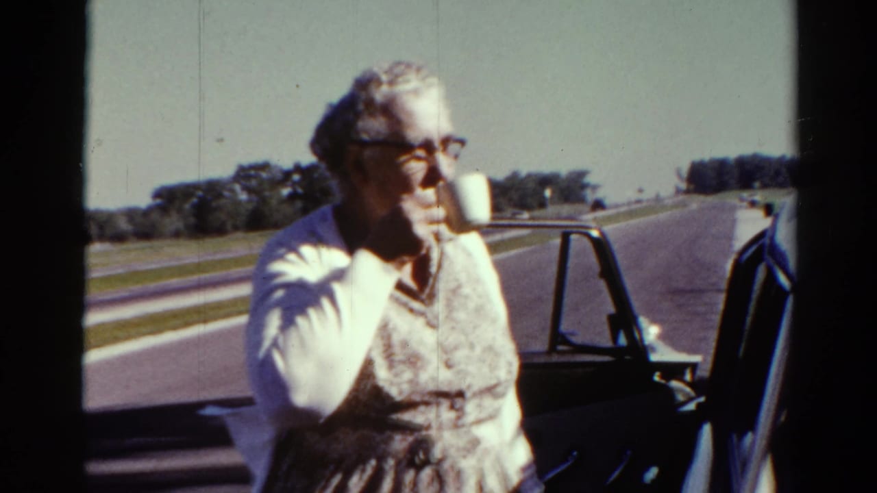 Older woman in a patterned dress and cardigan drinks from a white cup beside a car in Catskill, New York, 1962.