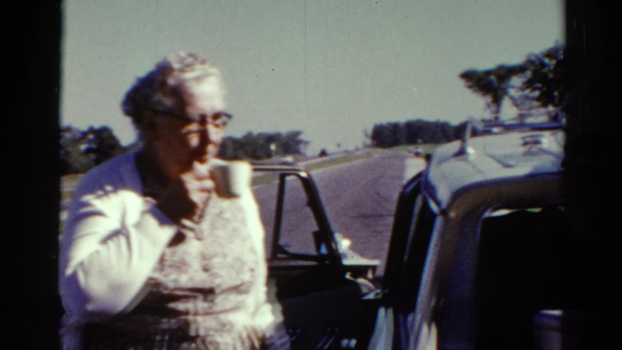 Older woman drinks from a cup near a vintage car during a sunny 1962 Catskill roadside stop.