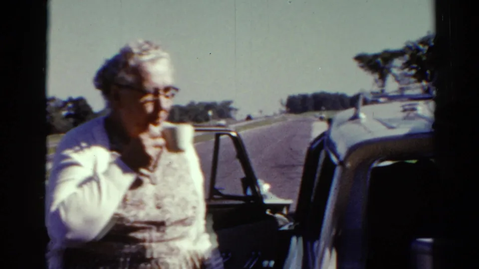 Older woman drinks from a cup near a vintage car during a sunny 1962 Catskill roadside stop.