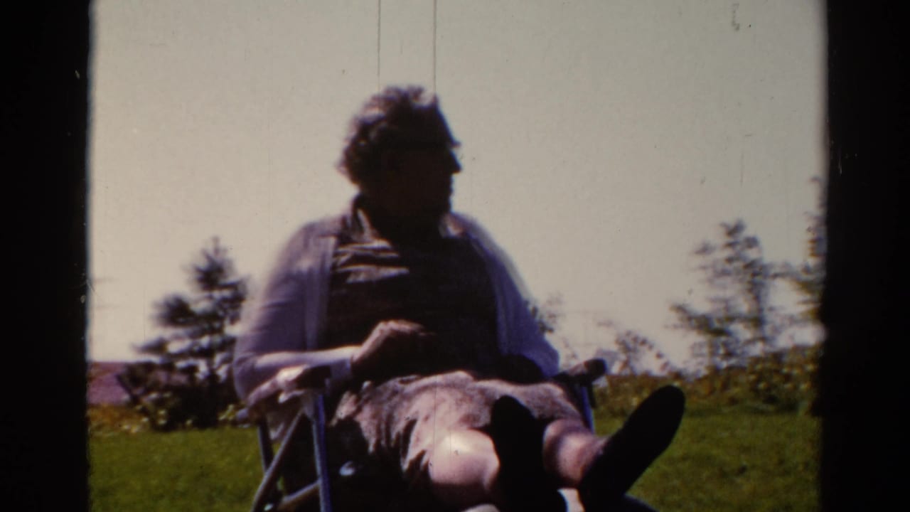 1962 Catskill home-movie frame of an adult man in glasses seated outdoors in a wheelchair or folding chair on a grassy slope.