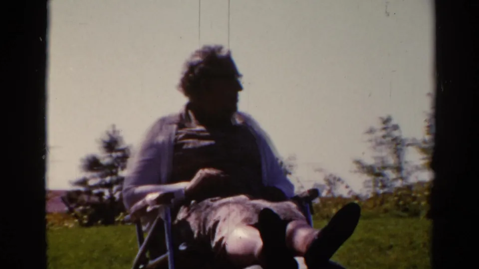 1962 Catskill home-movie frame of an adult man in glasses seated outdoors in a wheelchair or folding chair on a grassy slope.