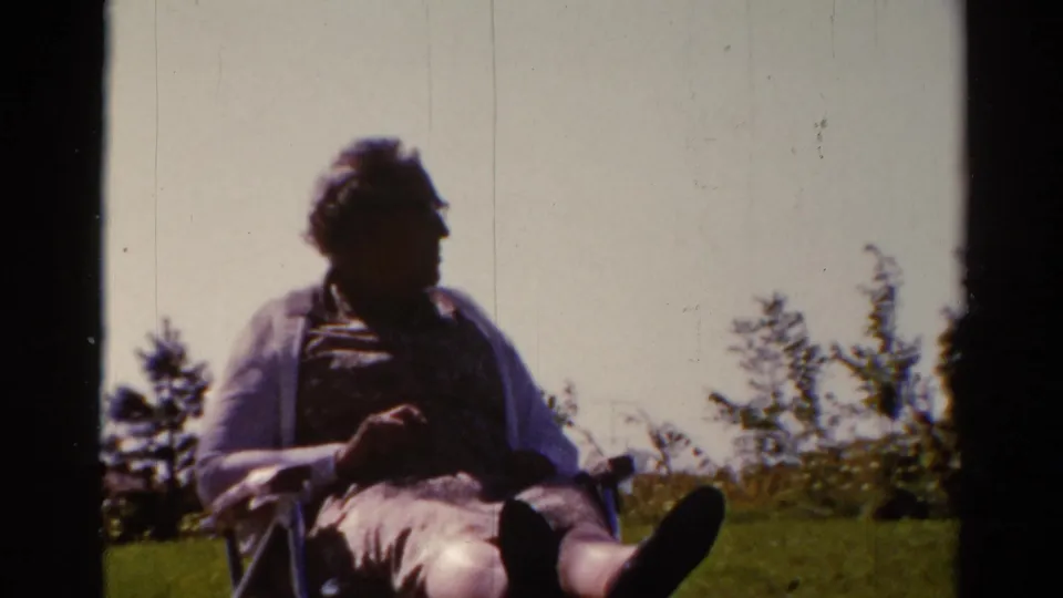 The adult man remains seated outdoors in the Catskill setting, with legs extended and trees behind him in soft 8mm color.