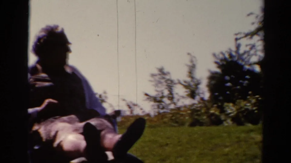 A 1962 Catskill home-movie frame partially crops the seated man at left, with grass, shrubs, and trees filling the view.