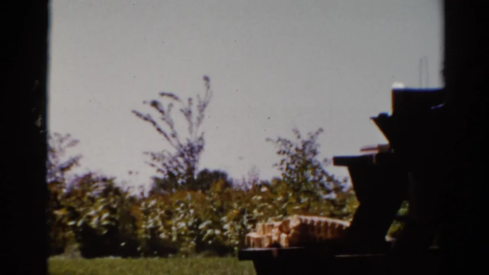 1962 Catskill yard view with a picnic table, stacked items, shrubs, pale sky, and dark foreground silhouettes at the frame edge.