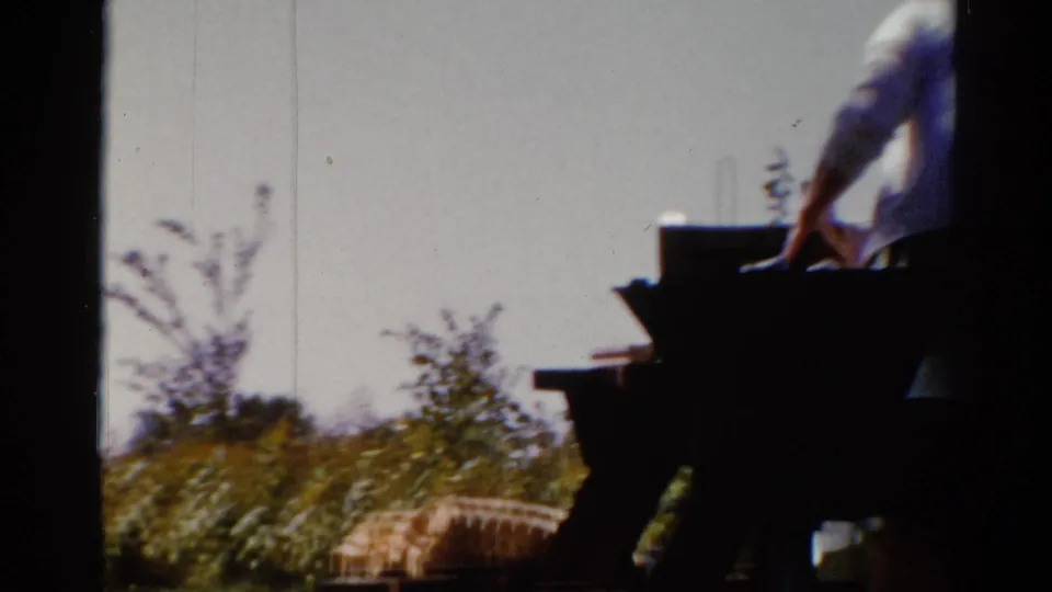 1962 Catskill home-movie frame with a partially visible adult leaning or moving near a picnic table in a grassy yard.