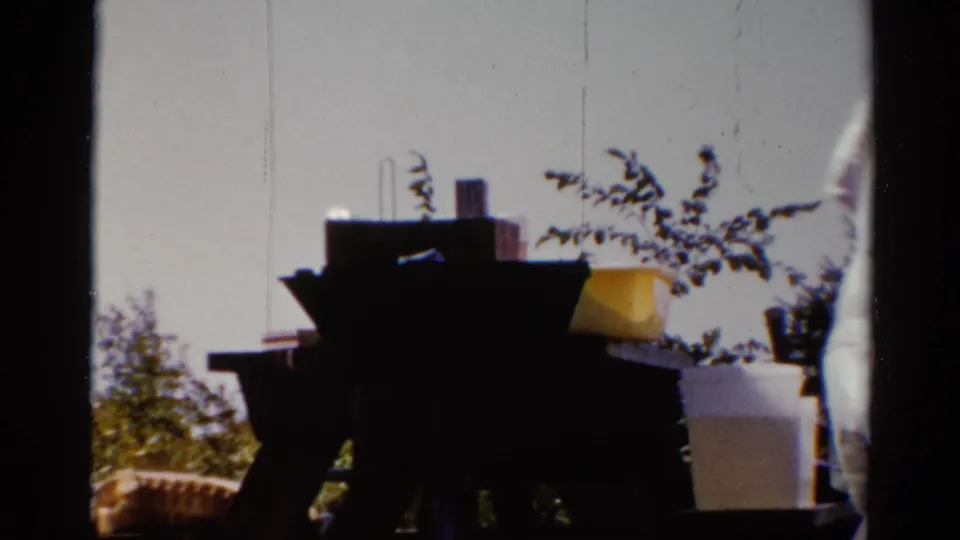 1962 Catskill yard scene with picnic table, containers or stacked items, shrubs, and a partially visible adult at the edge of frame.