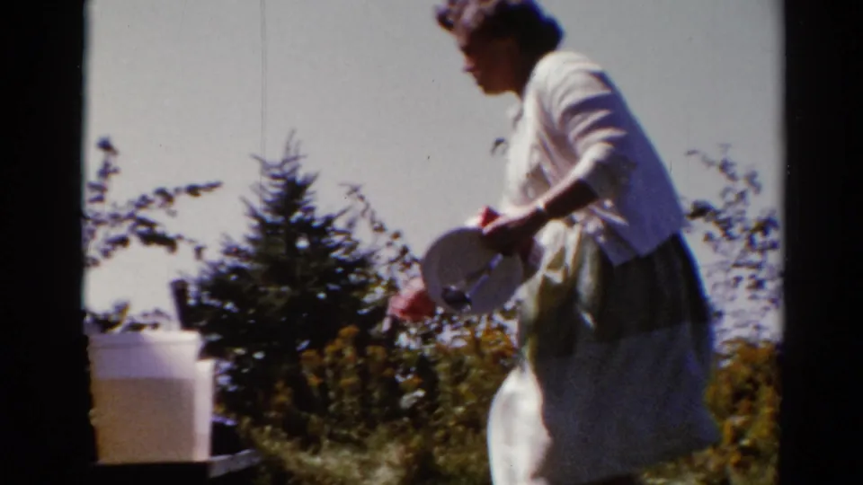 Adult woman in a white dress and light cardigan walks through a sunny Catskill outdoor scene, holding a round white object near a table.