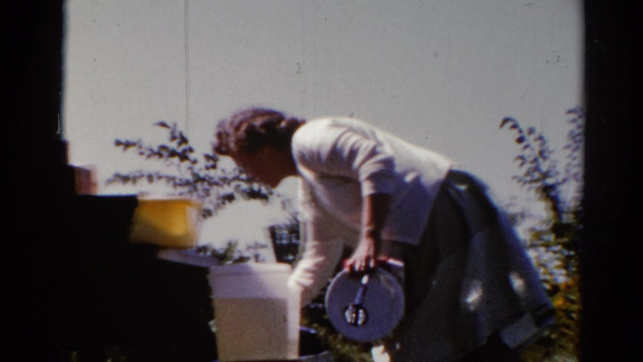 Woman in a white cardigan bends near outdoor dishes beside a backyard grill.