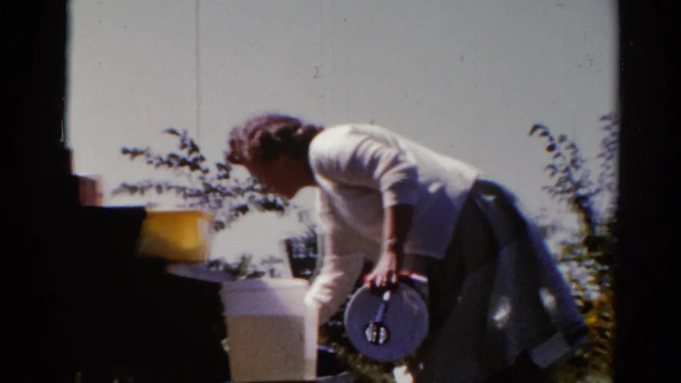 In Catskill, New York in 1962, a woman in a white cardigan bends near a plate or lid and outdoor cooking items in a sunlit yard.
