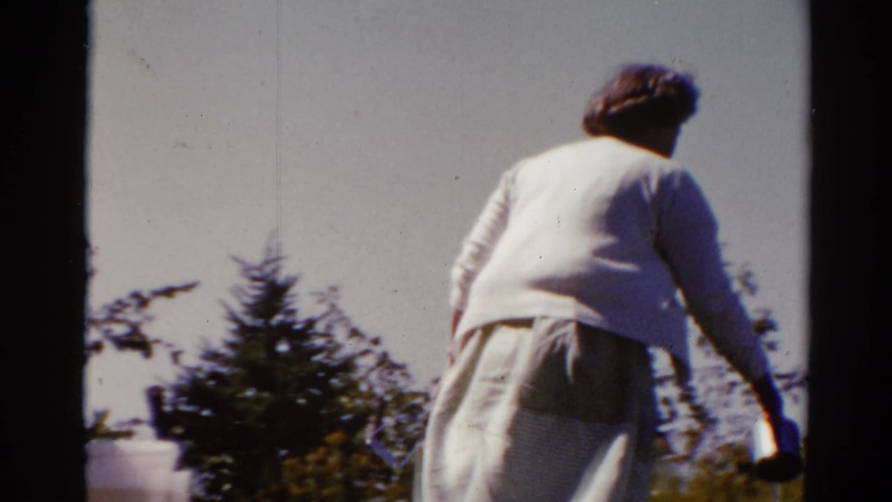 Woman walks away through a backyard carrying kitchen items near shrubs.