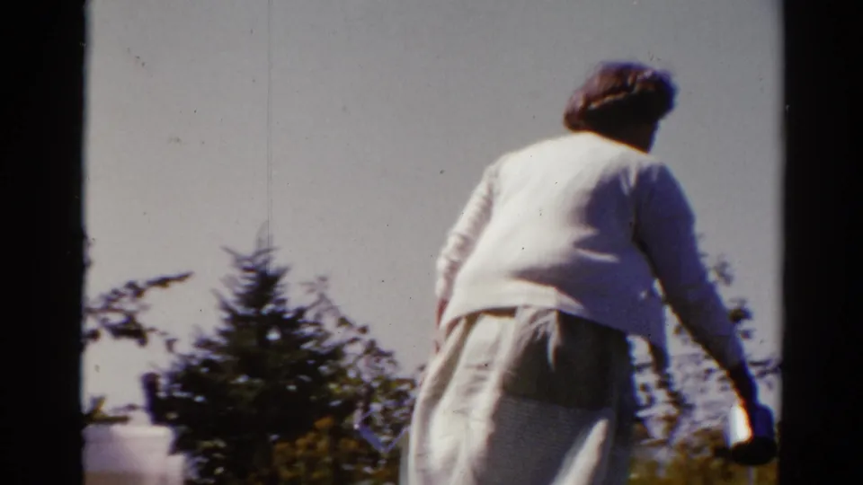 The woman, seen from behind in the 1962 Catskill backyard scene, carries kitchen items away from the cooking area.