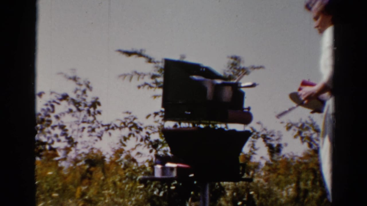 Woman in light clothing near a dark outdoor grill in a grassy area.