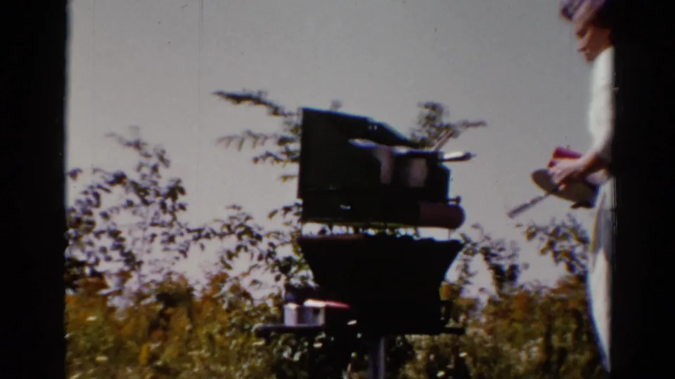 In 1962 Catskill home-movie footage, a woman in light summer clothing stands near a dark outdoor grill amid grass and shrubs.