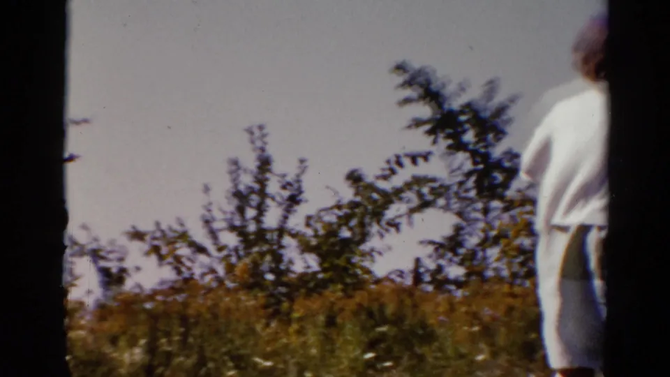 A soft, blurred frame shows the woman passing near a dark vertical frame edge, with grass and leafy shrubs behind her.