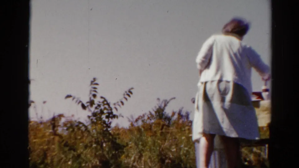 The woman is seen from behind beside a cloth-covered picnic table in a sunlit Catskill field clearing, 1962.