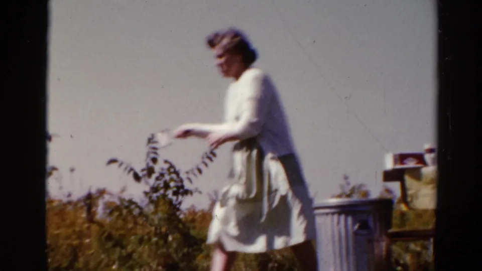 A woman reaches toward items on a picnic table near a metal trash can in the Catskill field clearing, 1962.