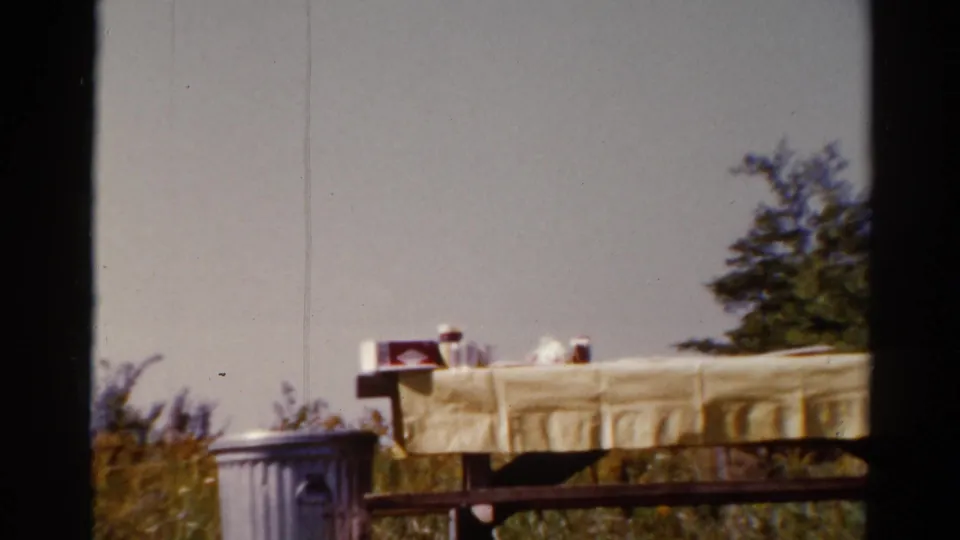 1962 Catskill home-movie frame showing a yellow-clothed picnic table beside a metal trash can and trees.