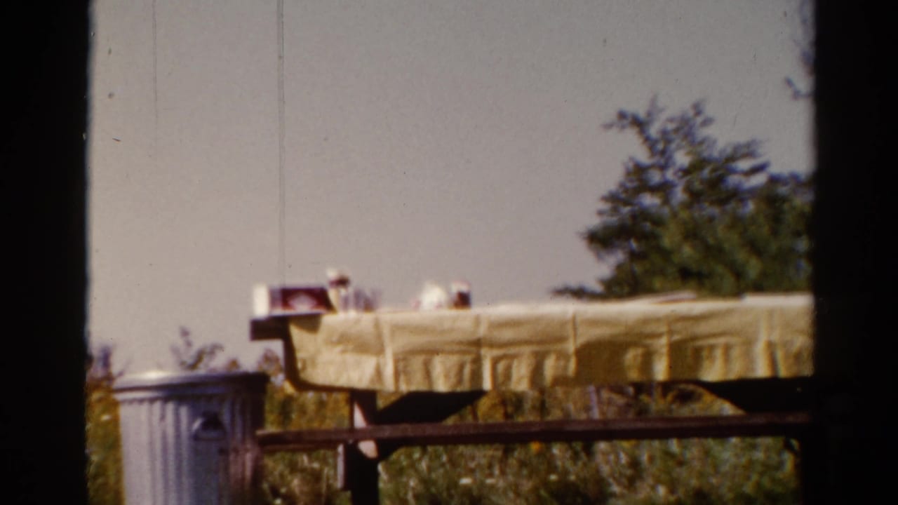 1962 Catskill frame with a yellow-covered picnic table, scattered small items, railing, and leafy trees.