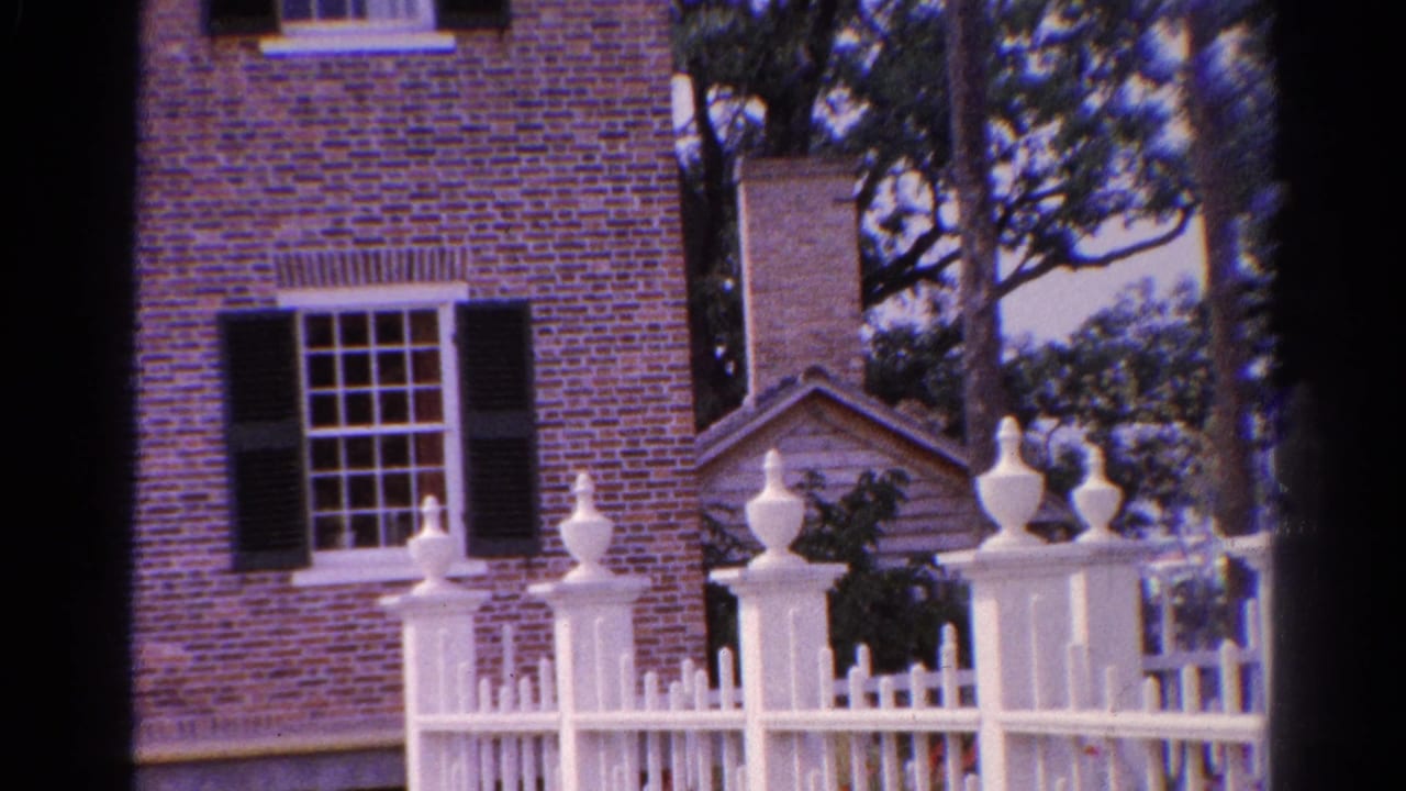 Catskill building exterior on 8mm film, with brick walls, black shutters, white fence finials, trees, and a small gabled entry detail.