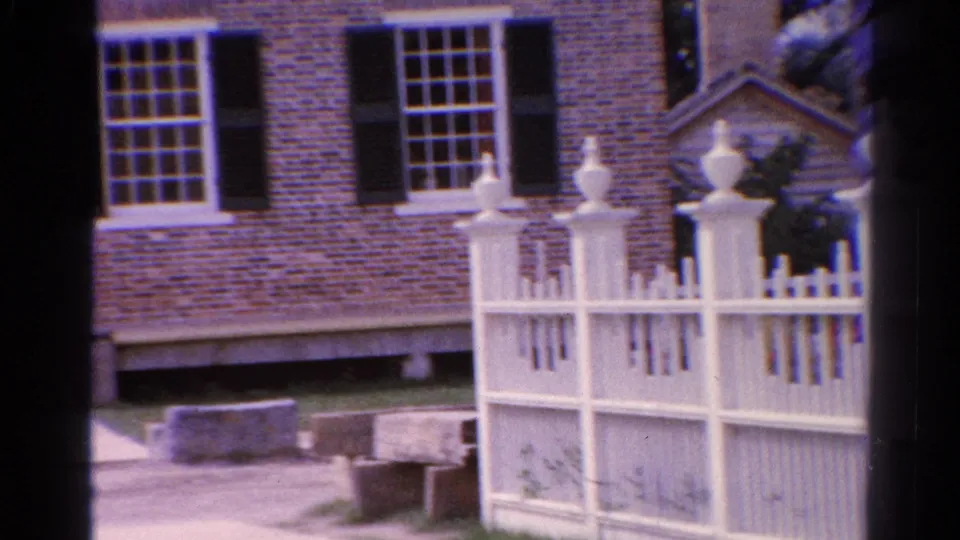 1962 home-movie frame of the Catskill brick building, showing lower windows, white picket fencing, finials, and part of the entrance area.