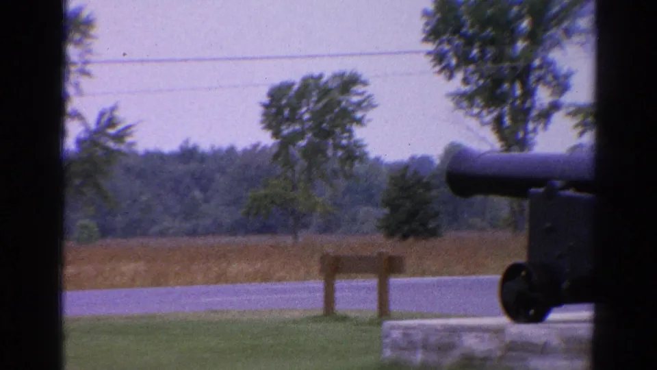 1962 Catskill, New York home-movie frame showing a dark cannon display near grass, trees, a road, and a small wooden signpost.
