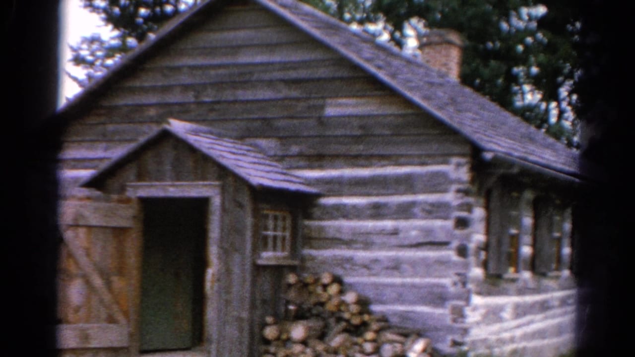 Weathered log cabin with open wooden door, stone base, chimney, and trees behind it.