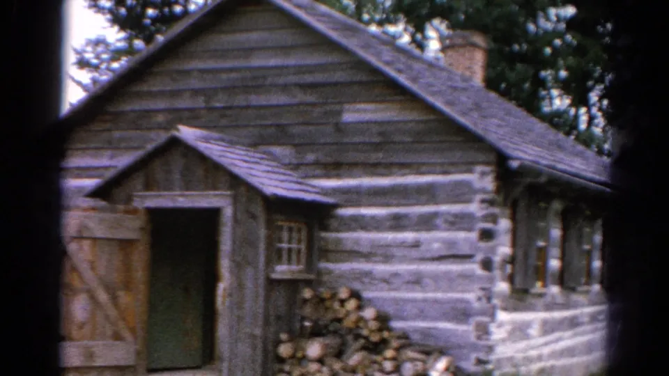 1962 Catskill-area 8mm frame of a rustic log cabin with an open door, stone foundation, small window, chimney, and leafy trees.