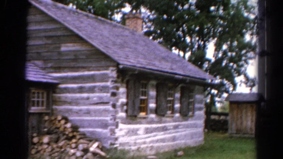1962 Catskill-area 8mm frame showing the side of a log cabin, stacked firewood, small windows, stonework, grass, and trees.