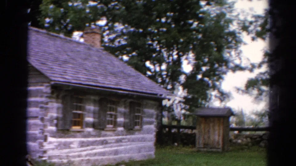 1962 Catskill-area 8mm frame of the cabin side with multiple windows, a shingled roof, trees, fencing, and a small wooden structure nearby.