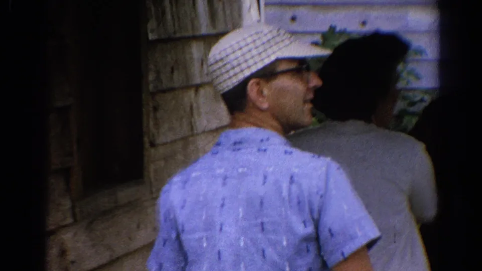In 1962 Catskill, New York footage, adults walk past a wooden-sided building, with a man in a checkered cap nearest the camera.