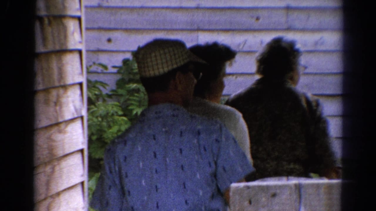 A small group walks away past wood siding and greenery, partly framed by dark film edges.