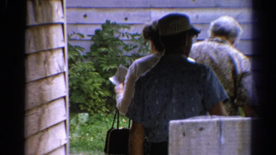 The handheld 8mm view follows adults from behind as they pass a wooden wall and move toward a green outdoor area in Catskill.