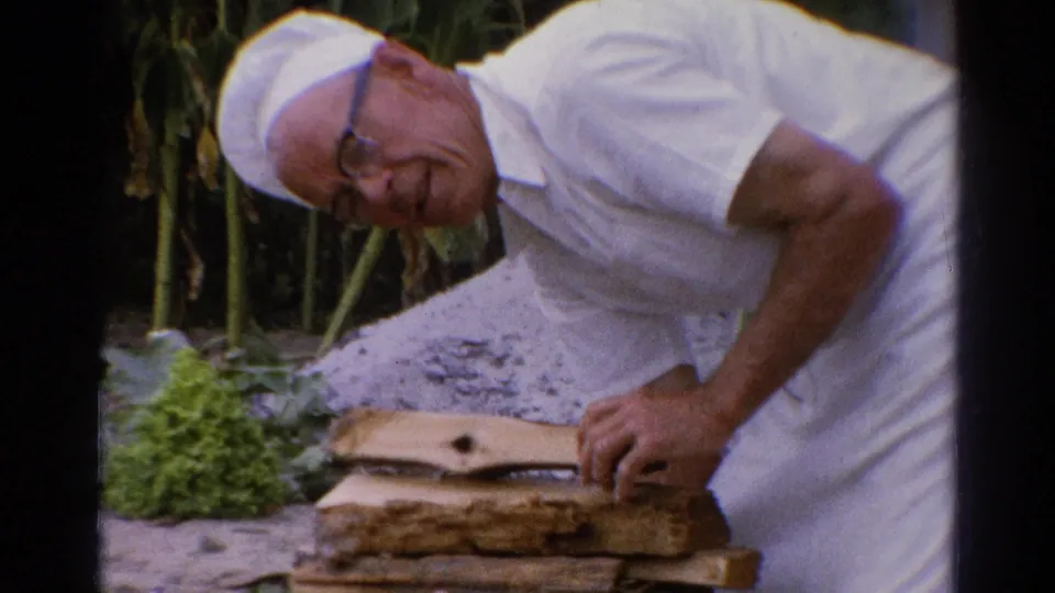 In 1962 Catskill home-movie footage, an older man in white clothing and cap bends over stacked firewood near leafy garden plants.