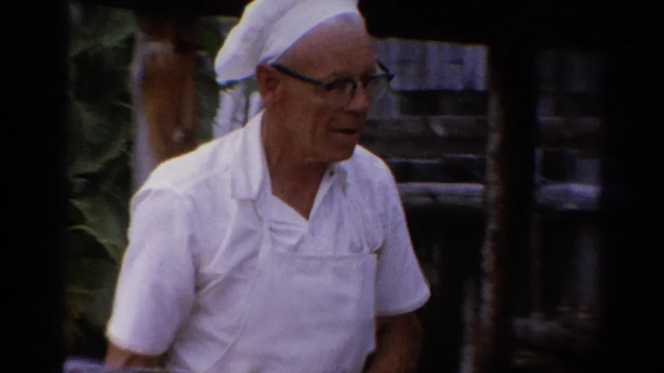 An older man in white work clothing and glasses stands in partial profile in the 1962 Catskill garden footage.