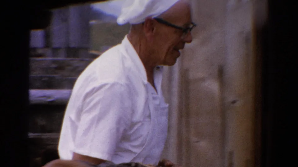 The man in white clothing and cap moves past a light-colored outdoor wall, with garden surroundings partly visible.