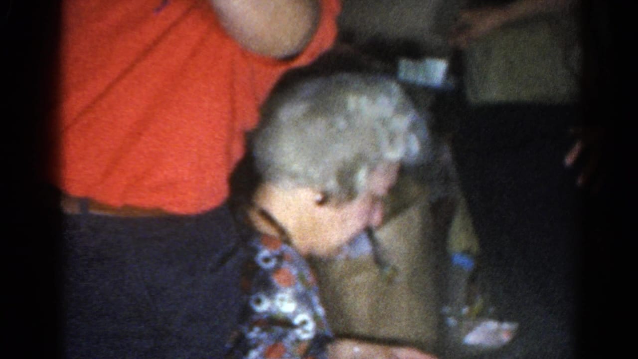 Elderly woman with white hair leans forward beside a person in a red shirt in a dim room.