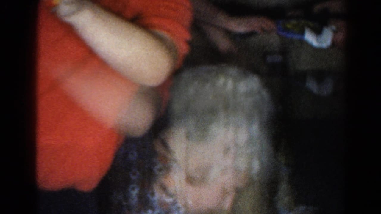 A soft 8mm frame shows the elderly woman bending forward during the holiday gathering, with a red-shirted adult partly visible above her.