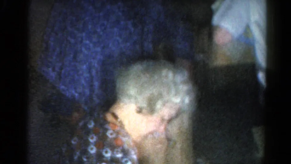 The elderly woman remains seated or bent forward while relatives in patterned clothing stand close in the dim Catskill interior.