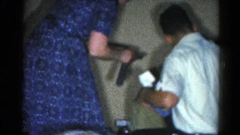 A blue-dressed adult stands near a seated man in a white shirt who appears to handle a small gift or paper in the private holiday room.