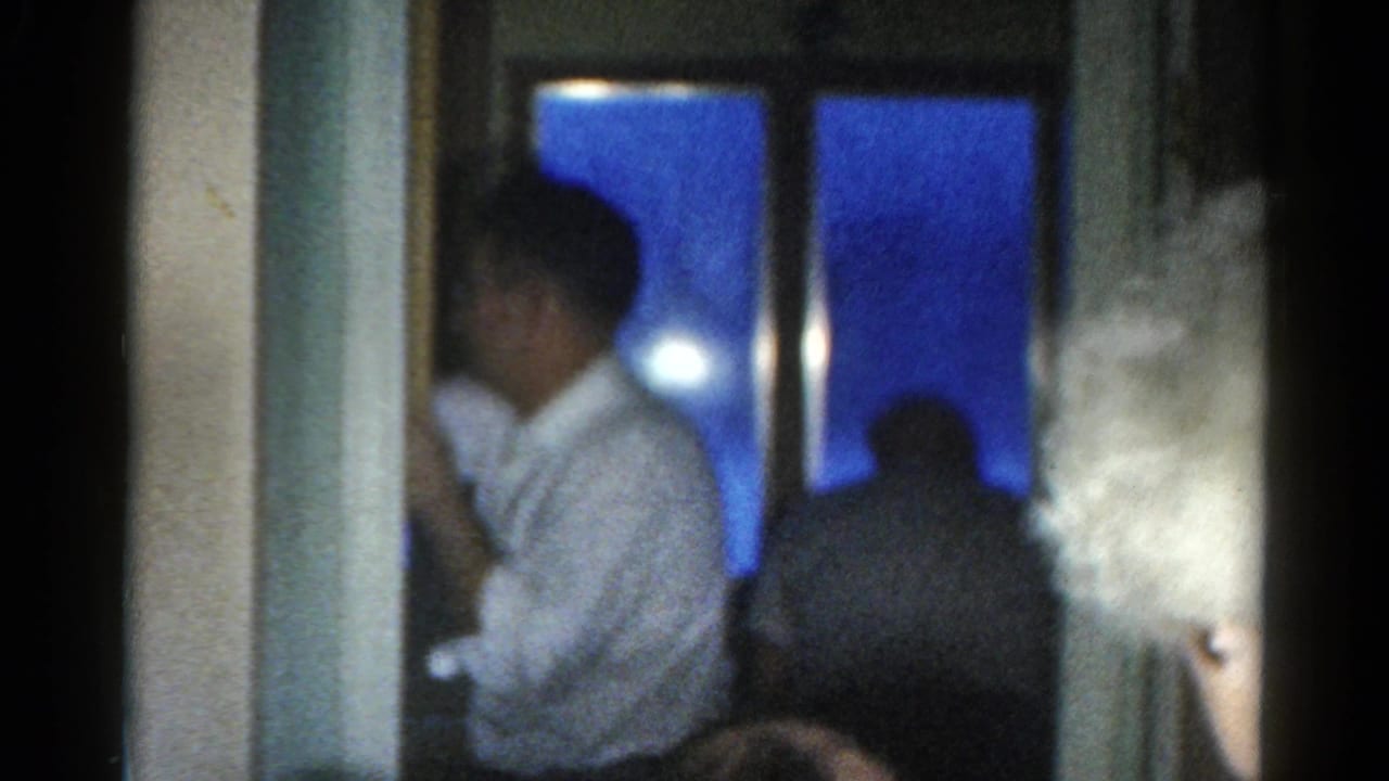Dim Catskill interior with a seated man and another figure near a blue night window, filmed on 8mm in 1962.