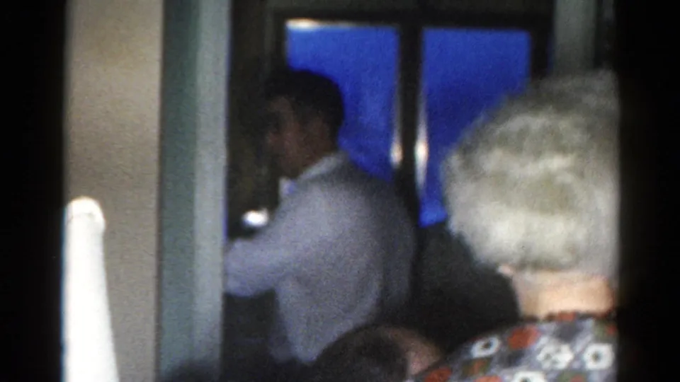 A gray-haired adult in a patterned blouse moves into the foreground of a dim 1962 Catskill home interior.