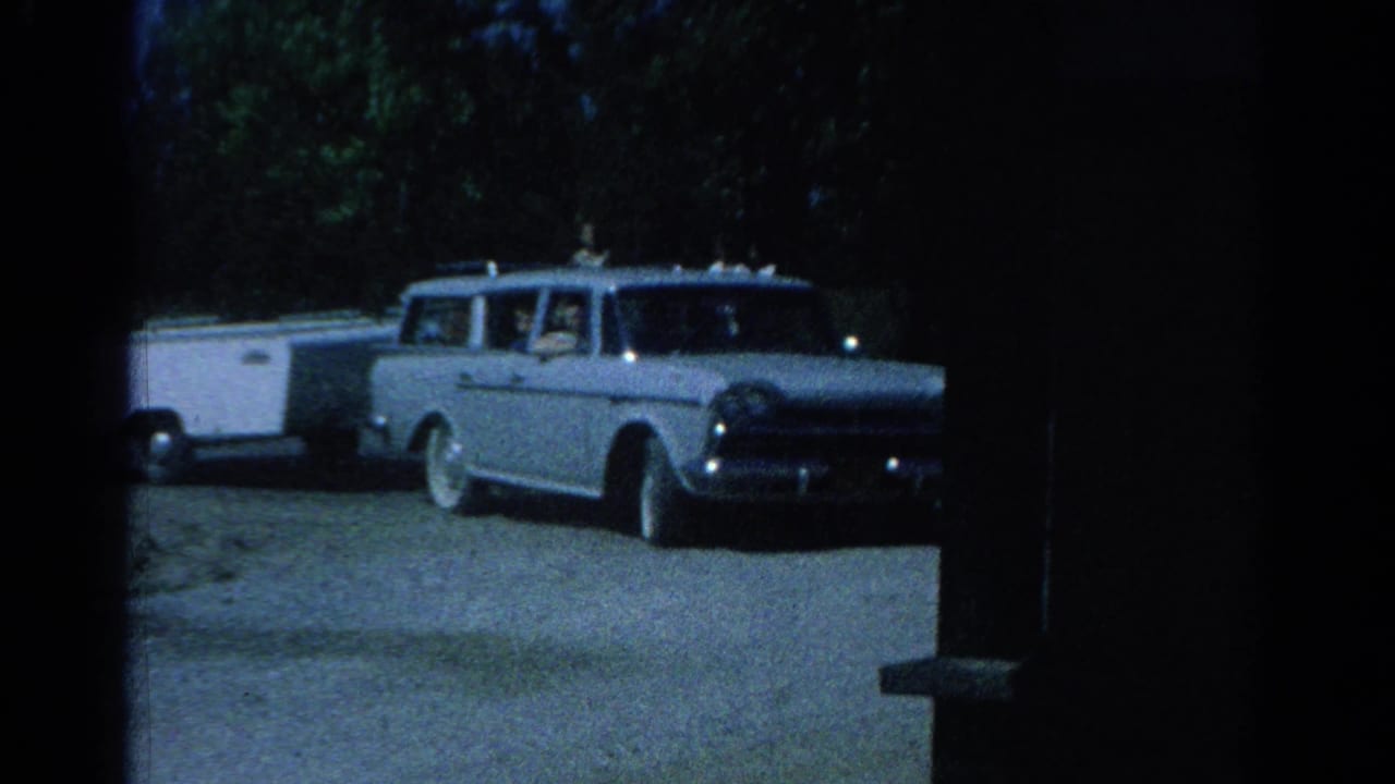 1962 Catskill home-movie frame showing a blue station wagon pulling a white camper trailer through a wooded roadside area.