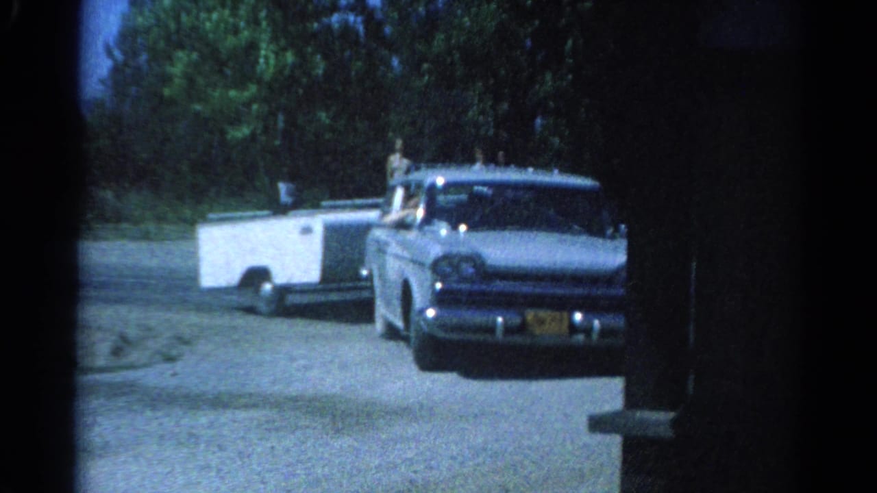 1962 Catskill frame of a blue station wagon towing a white camper, with people visible in or near the vehicle roof area.