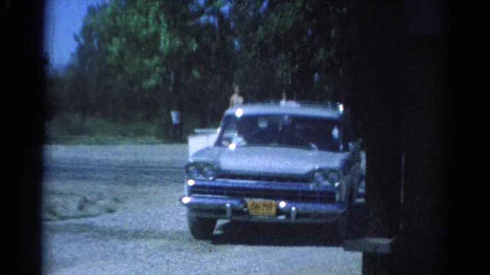 The blue station wagon faces the camera in Catskill-area 1962 footage, towing a small white camper past trees and gravel.