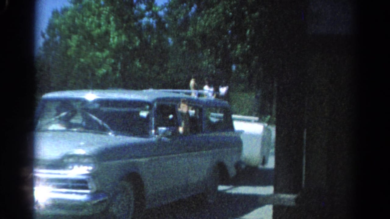 1962 Catskill home-movie frame showing a light station wagon pulling a white camper trailer past trees and a low roadside wall.