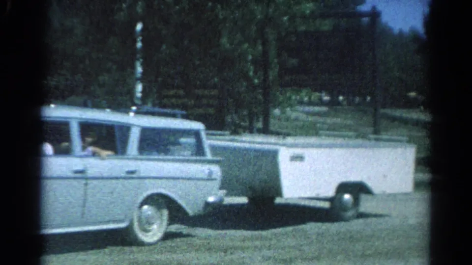 A light station wagon tows a white camper trailer along a tree-lined Catskill roadside, with a signboard visible in the background.