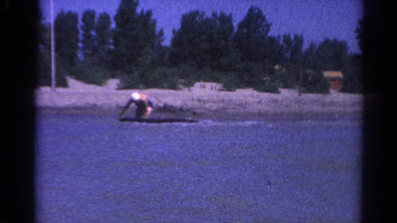 Woman in a white swim cap crouches on a small board in shallow lake water near a sandy tree-lined shore.