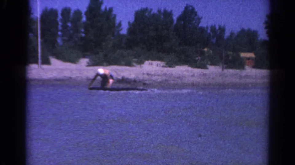 In Catskill, New York, 1962, a woman in a white swim cap appears to balance low on a small board in shallow lake water.